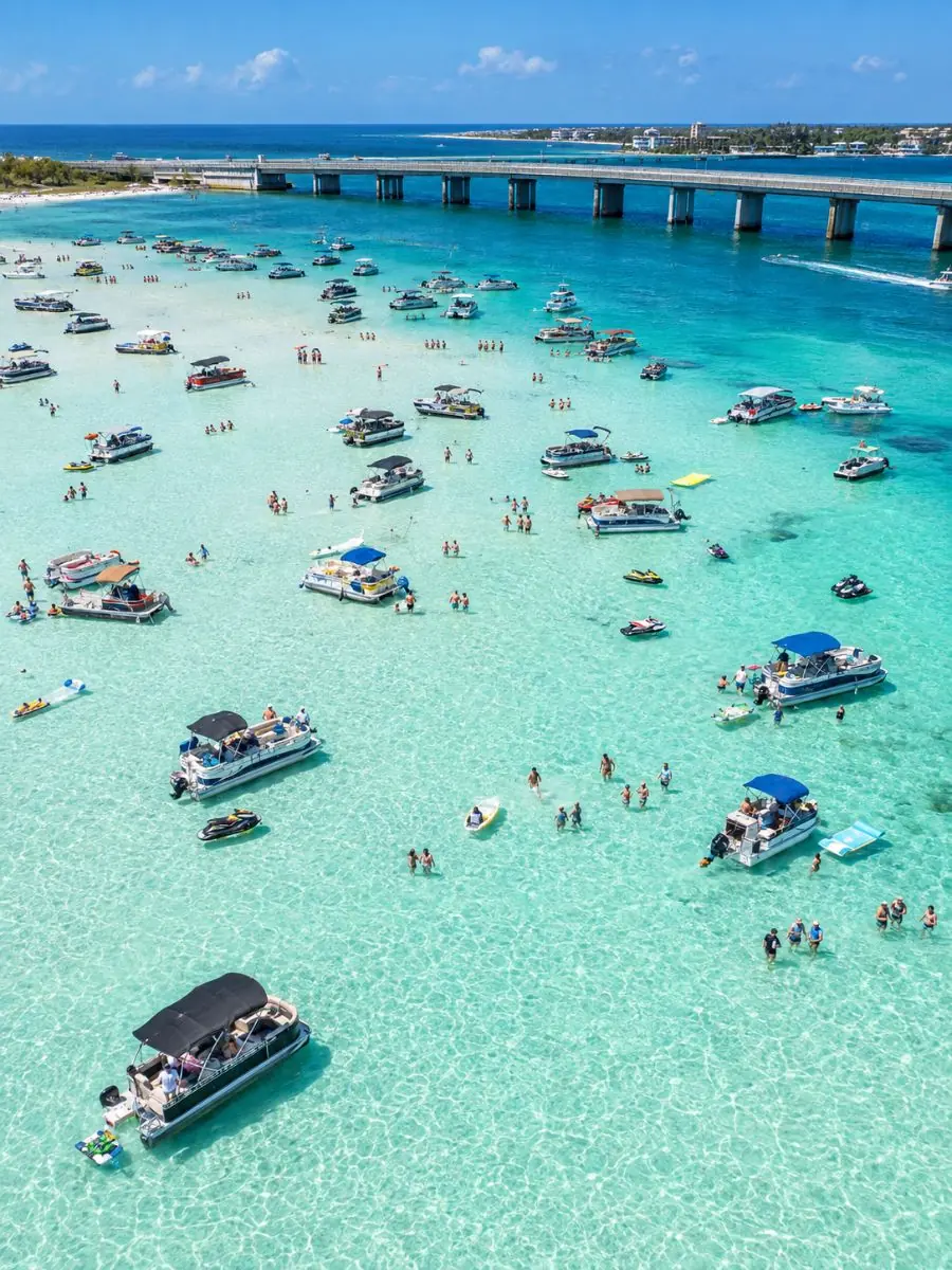 Luftaufnahme von Booten und Jetskis, die in türkisfarbenem Flachwasser über einer Sandbank nahe einer niedrigen Brücke ankern