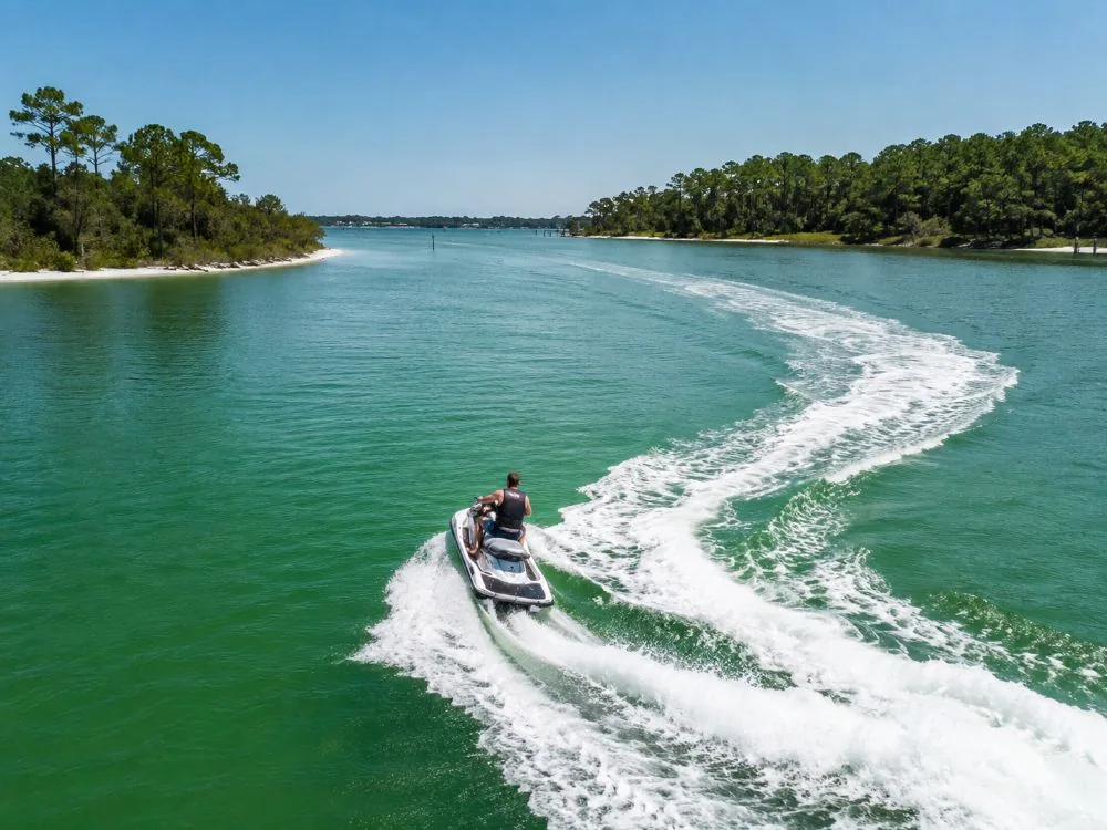 Ein Jetski zieht eine lange, geschwungene Heckwelle über eine weite, ruhige Bucht mit flachen, bewaldeten Ufern