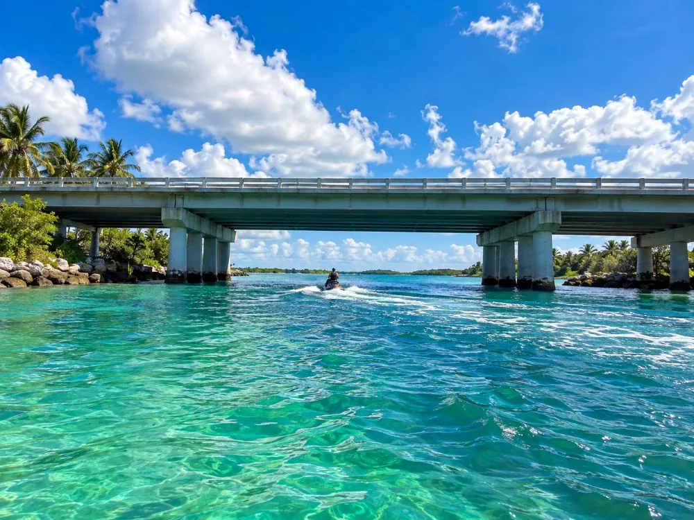 Eine lange, niedrige Betonbrücke über einem grünen Gezeitenkanal, weit darunter ein Jetski