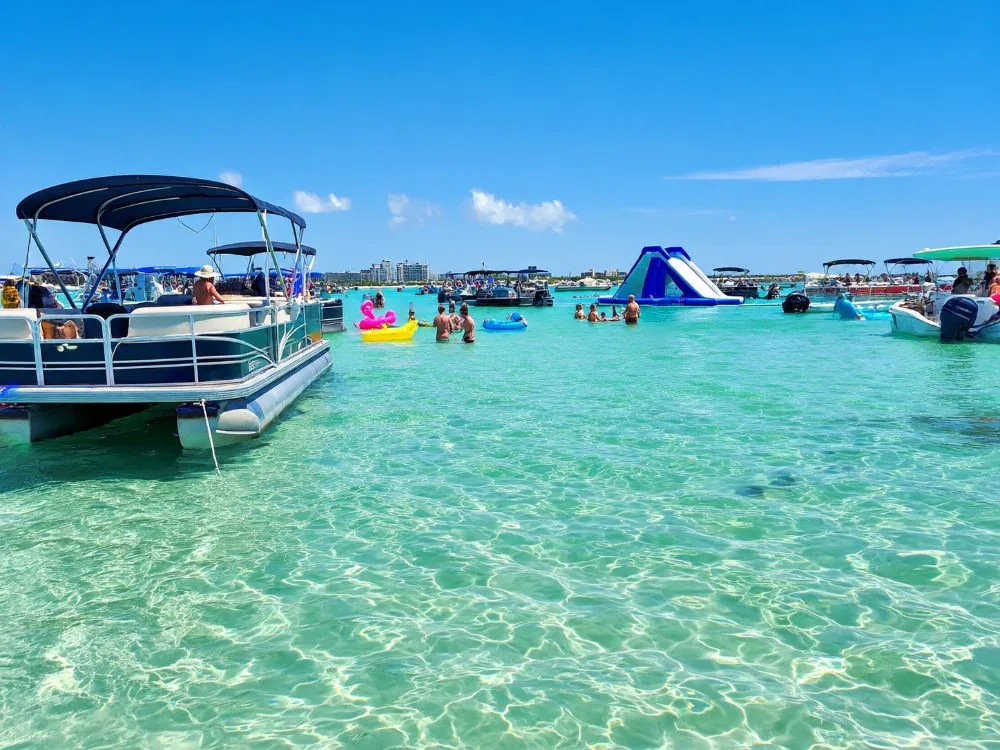 Boote ankern in knietiefem türkisfarbenem Wasser über einer Sandbank, mit Schwimmreifen und einer Wasserrutsche