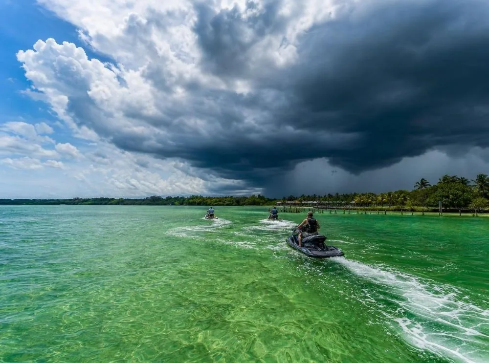 Dunkle Gewitterwolken türmen sich über einer sonnigen Bucht auf, während Jetskis zu einem Steg zurückfahren