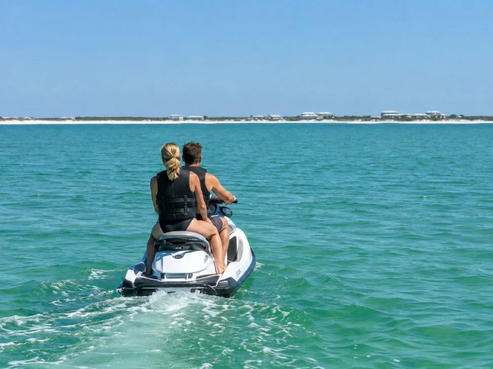 Zwei Erwachsene in Schwimmwesten fahren langsam auf einem Jetski über ruhiges smaragdgrünes Wasser, von hinten gesehen